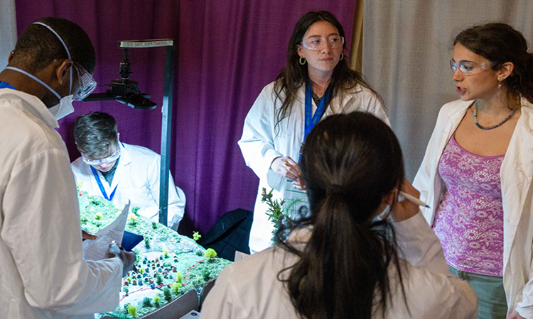 Michigan Tech students in lab coats and safety goggles discuss their bench-scale demo.